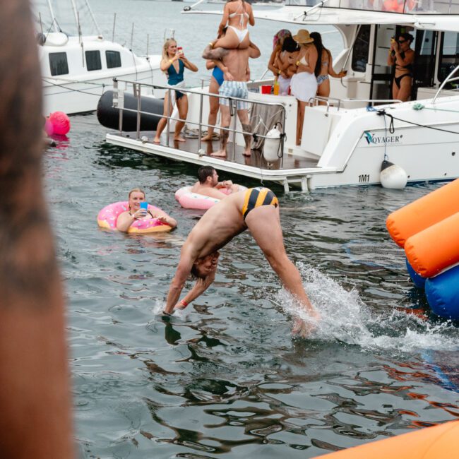 A man dives into the water from the edge of a boat, wearing a black and yellow striped swimsuit. In the background, people socialize on a yacht and in the water. An inflatable flamingo float and orange buoy are also visible nearby.