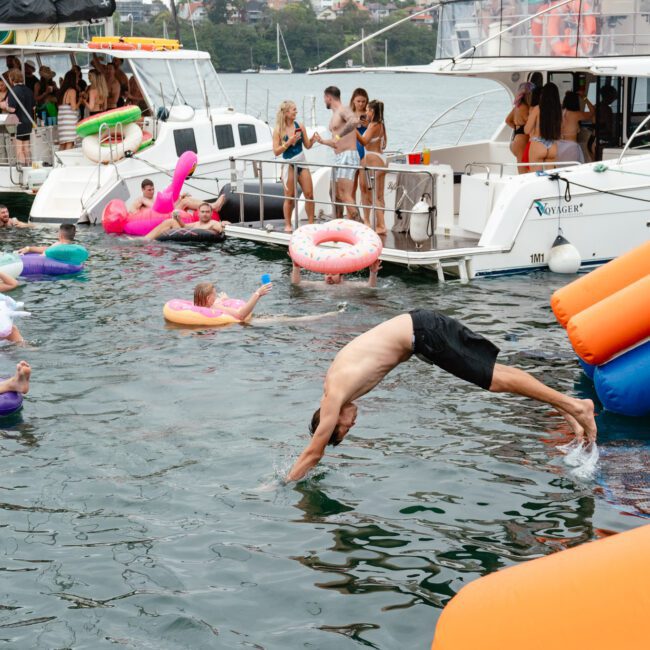 A crowded summer water party on a lake with two boats docked nearby. People are swimming and floating on inflatables. A man is mid-dive off an orange and blue slide. Onboard, more party-goers are mingling, taking photos with exuberant smiles. Trees and buildings provide a picturesque backdrop in the background.