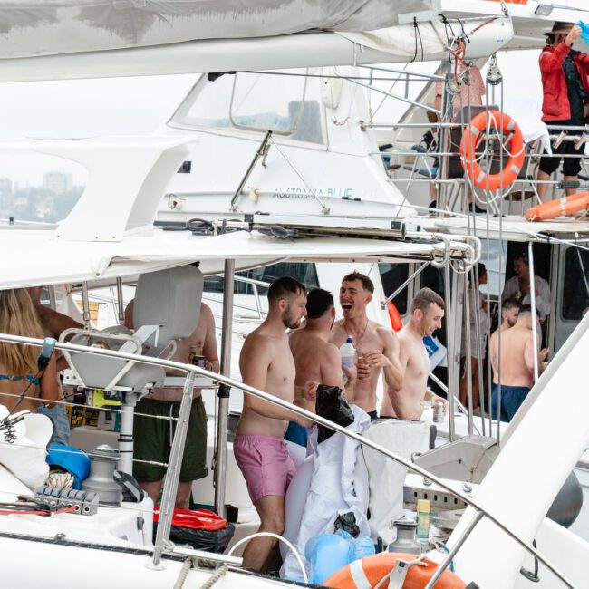 A lively scene on a yacht features a mix of men and women enjoying a party. Several people are shirtless, holding drinks, and engaging in conversation. The yacht is docked with life jackets visible against the backdrop of a foggy city skyline, adding to the festive atmosphere.