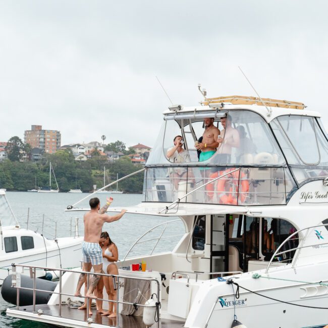 A group of people enjoying a party on a docked boat. Some are on the upper deck under a canopy, waving and taking pictures, while others are on the lower deck or swimming nearby. Several buildings and lush trees are visible in the background under a cloudy sky.