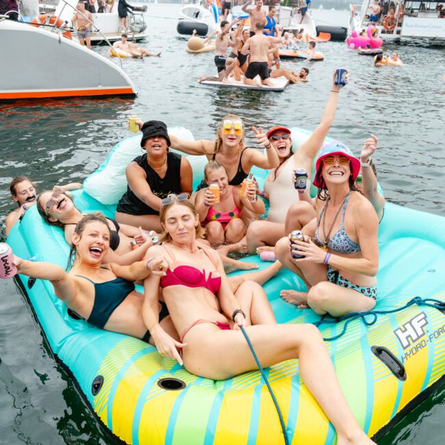 A group of smiling friends in swimwear sit on an inflatable floatie in the water, holding drinks and posing for the camera. Other people are visible laughing and relaxing in the background on boats and inflatables, creating a lively and festive beach atmosphere.