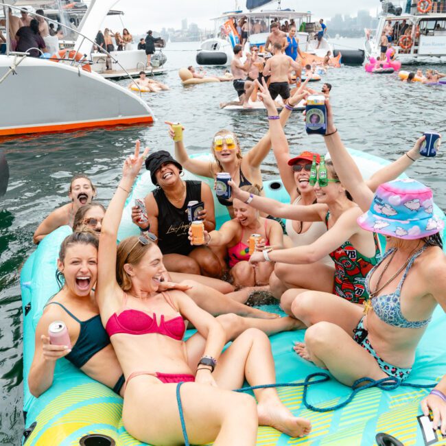 A group of people in swimsuits enjoy a lively gathering on an inflatable raft in the water. They are laughing, holding drinks, and posing with their arms raised in celebration. Surrounding them are other rafts and people having fun in the water, with boats sailing in the background.