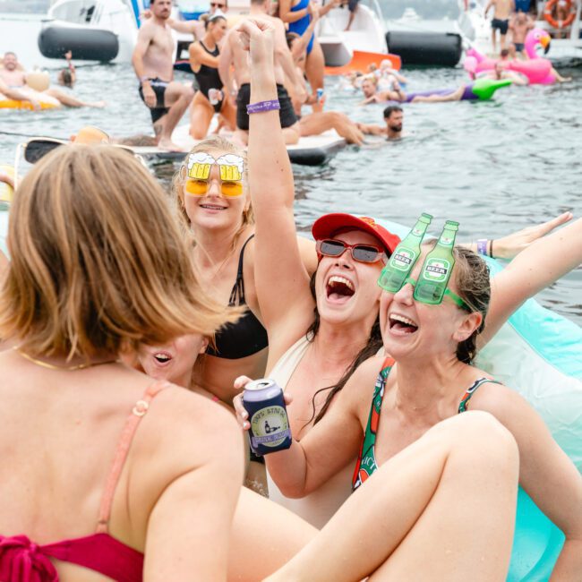 A group of people in swimsuits celebrates joyfully on inflatable floats in the water, raising drinks and laughing while wearing sunglasses. They are surrounded by others also enjoying the sunny day and relaxing on floats.