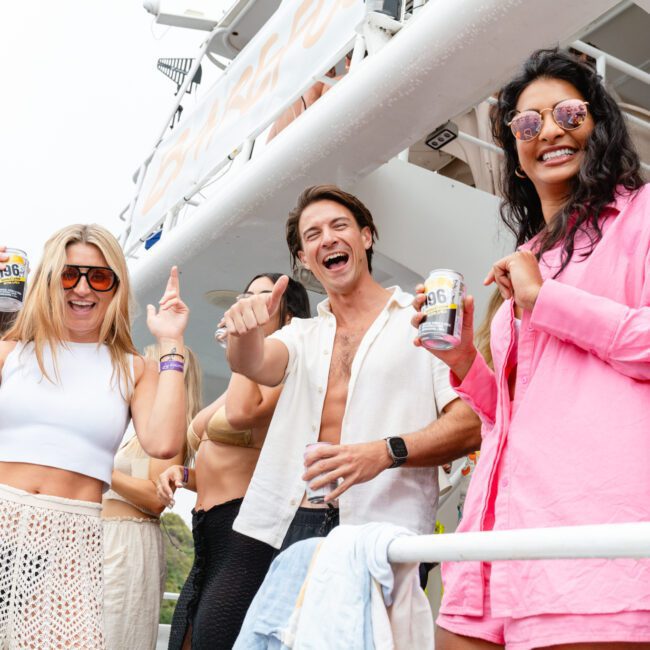 A group of people enjoying themselves on a boat, holding canned beverages and wearing casual summer attire, including shorts, swimsuits, and sunglasses. Everyone is smiling and having a good time with some delicious snacks. The setting appears to be festive and lively.