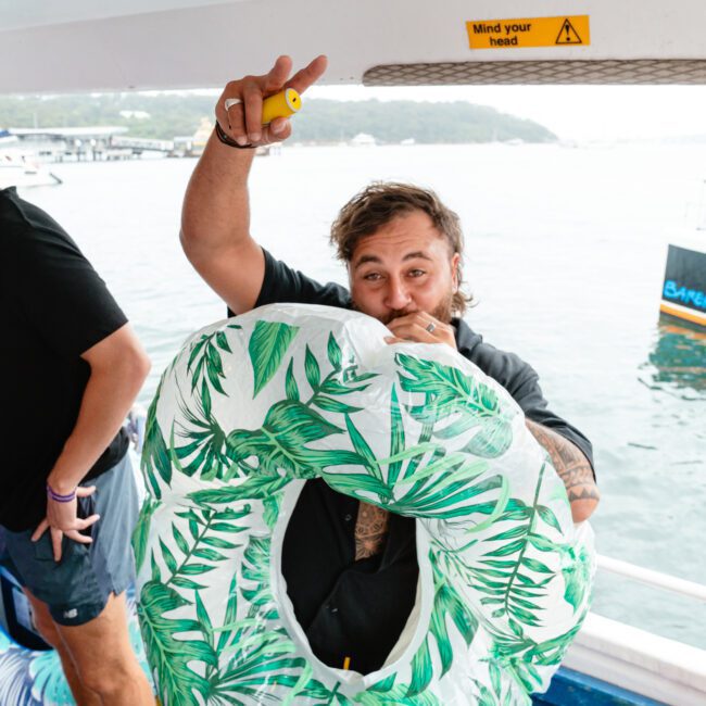 Two people are on a boat, with one man in the foreground holding a tropical leaf-patterned inflatable ring, playfully pointing and smiling. The other man in the background is wearing a black shirt. A "Mind your head" sign is visible above them, with water and boats creating a lively backdrop.