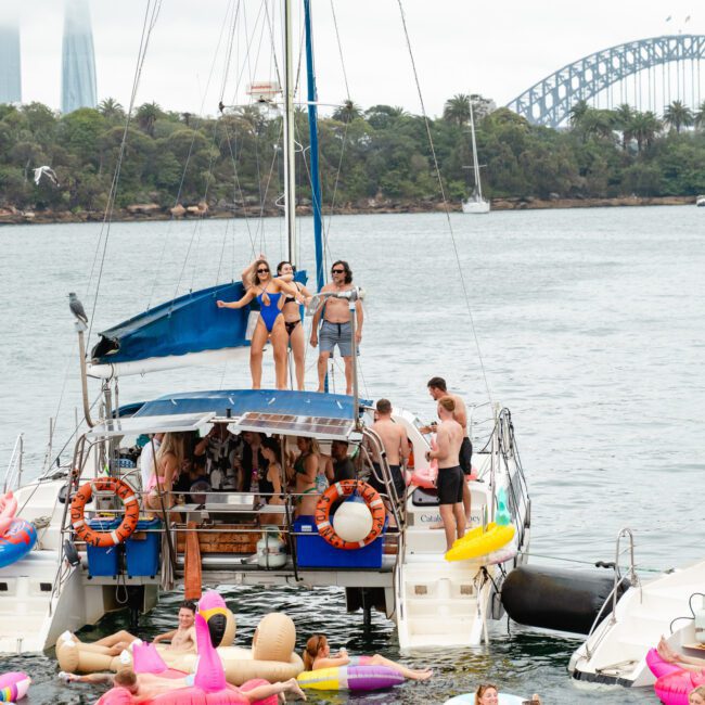 A lively boat party is taking place on a catamaran with numerous people enjoying the festivities. Some attendees are on the boat, while others swim and lounge on inflatables in the water. In the background, a stunning city skyline and an elegant arched bridge set the scene.
