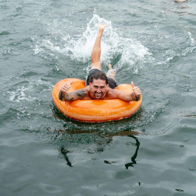 A man smiles and gives a thumbs-up while lying on an orange pool float in the water. Other people can be seen swimming and floating in the refreshing, sunlit background.