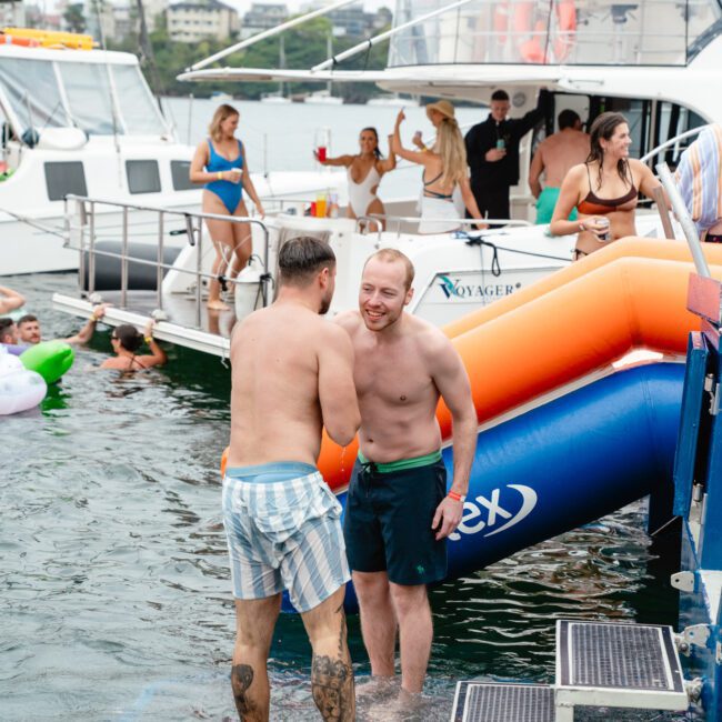 Two men, shirtless and in swimsuits, stand on a floating platform in the water, smiling and shaking hands. Behind them is an inflatable orange slide leading from a docked boat. Several people are on the boat and in the water, enjoying a vibrant atmosphere with laughter and fun games.