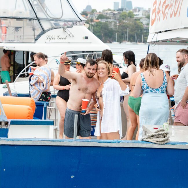 A group of people celebrating on a boat, with many holding drinks and socializing. In the foreground, a man and a woman pose for the camera. Other boats and people are seen in the background, with a stunning waterfront cityscape in the distance.