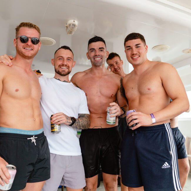 A group of five shirtless men, some holding drinks, are smiling and posing together on a boat under the bright sun. The background showcases parts of the boat's sleek interior and exterior. They appear to be having a fantastic day out on the water.