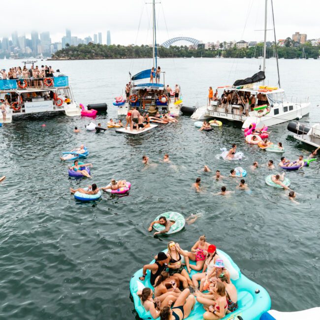 A lively gathering on a harbor with several boats and yachts. People swim and float on inflatable rafts, paddleboards, and inner tubes, enjoying the water. The harbor is surrounded by a city skyline in the background, including an iconic bridge peeking through the mist.