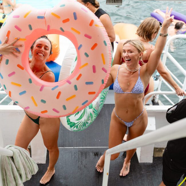 Two people in swimsuits are on a boat's deck, smiling and posing. One holds a pink, donut-shaped pool float. Others are in the water nearby with more floats. The scene is vibrant and joyful, capturing a sensational summer day of fun and swimming.