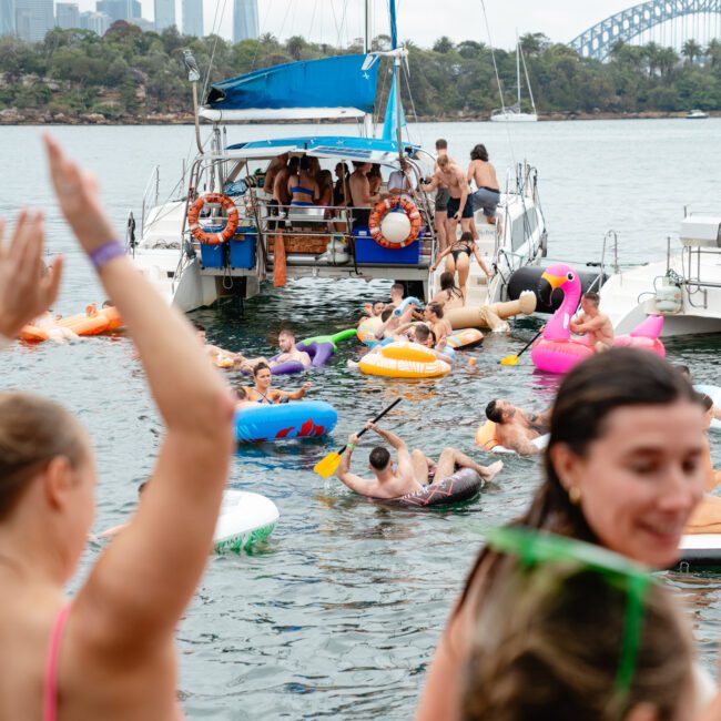 A group of people enjoying a vibrant floating party on a body of water, surrounded by various inflatable floats, including a flamingo. Some are on a boat in the background, with a bridge and cityscape visible in the distance. Two women are in the foreground, one with her arms raised joyfully.