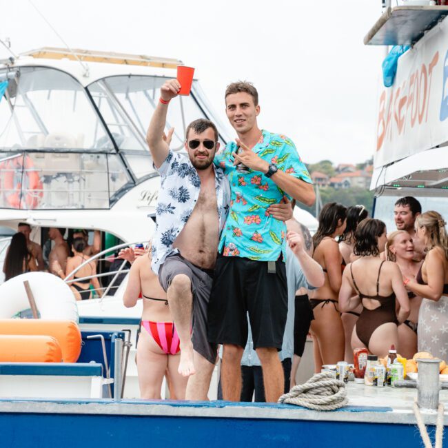 Two men in tropical shirts stand on the deck of a boat. One lifts a red cup and has his arm around the other, who holds up a peace sign. Behind them, many people in swimwear socialize and enjoy the lively boat party. In the background are more boats and shimmering water.