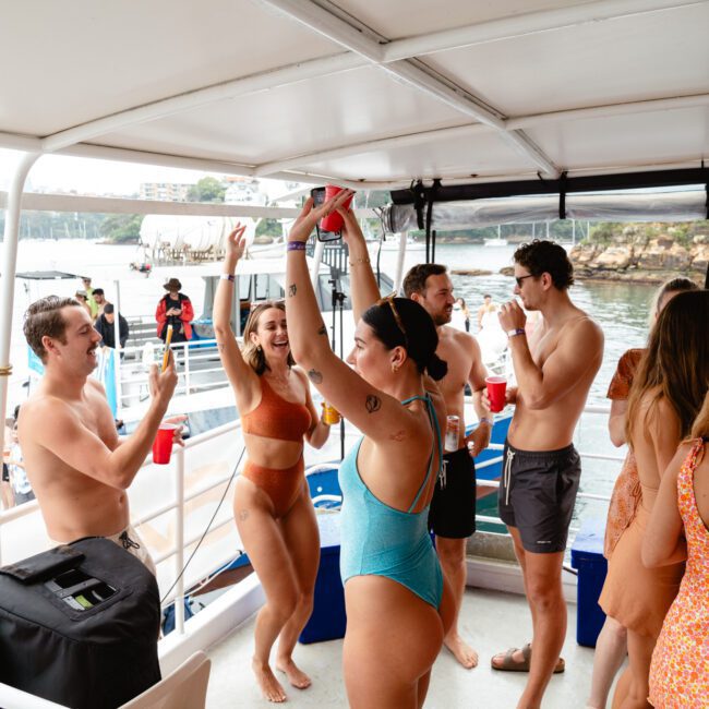 A group of people in swimwear is gathered on a boat, dancing and holding drinks. They are smiling and appear to be enjoying a sunny day on the water. The background shows water, docks, and other boats. The atmosphere is lively and vibrant, perfect for a summertime celebration.