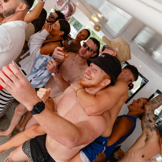 A lively group of people are partying on a boat, holding drinks and smiling broadly. Participants are dancing and engaging enthusiastically, with one person giving another a piggyback ride. The background shows parts of the boat's vibrant interior, adding to the festive atmosphere.