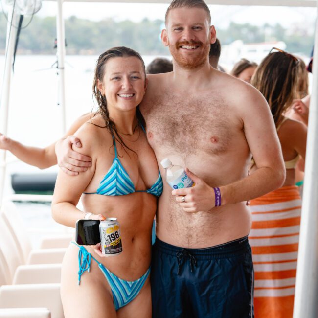 A smiling man and woman in swimwear pose together on a boat. The woman, in a striped bikini, holds a drink, while the man, in black swim trunks, holds a beverage can and water bottle. People and clear blue water are visible in the background.
