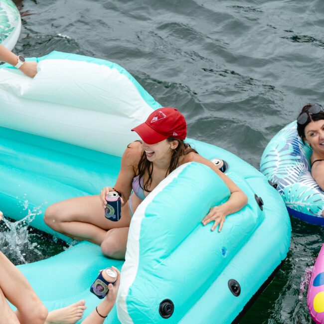 Four people are floating on inflatable pool floats in a serene body of water. One person in a red hat and sunglasses holds a canned drink, smiling. Others are relaxing and chatting on vibrant, colorful floats. Legs and feet of another person are visible in the foreground, adding to the cheerful scene.