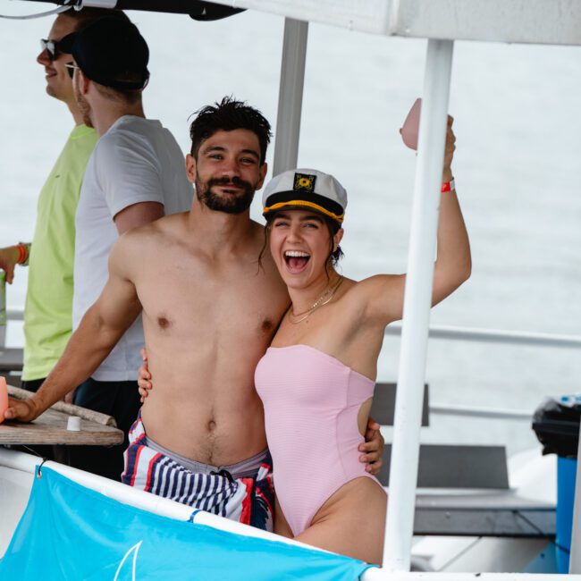 A man and woman pose happily on a boat. The man, shirtless in striped shorts, has his arm around the woman. She wears a pink swimsuit and a captain's hat, smiling while holding up a drink. The couple enjoys their time as other people relax in the background on the boat.