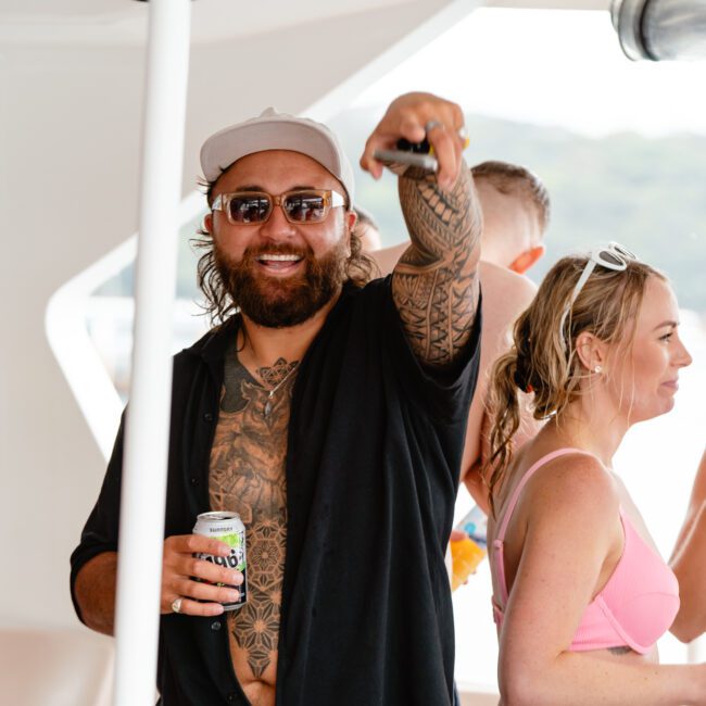 A bearded man with tattoos, wearing sunglasses and a cap, holds a drink and points towards the camera on a boat. He is surrounded by other people, some in swimwear, enjoying drinks and socializing. A Bluetooth speaker plays music in the background, with a blurred view of water and greenery adding to the relaxed vibe.