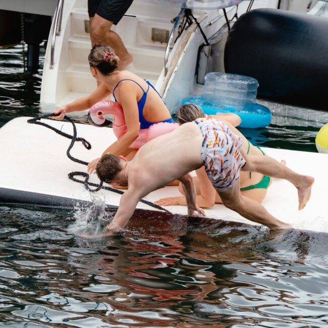 Two people are diving off a floating platform into the water, while two others sit on the edge. One person is holding an inflatable unicorn ring. The scene is set on a boat dock with paddleboards and various colorful water toys in sight.