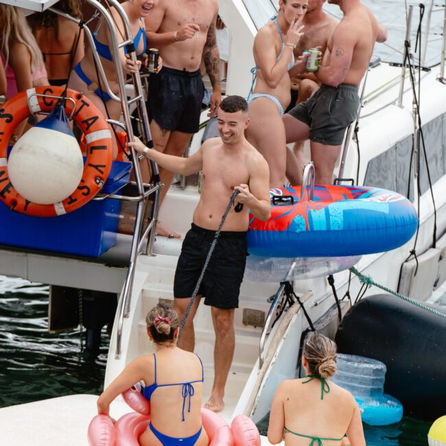 A group of people in swimsuits is enjoying a lively party on a boat. A shirtless man with a microphone and an inflatable ring stands confidently on a platform between two boats, while others stand or sit nearby. Colorful inflatable rings and a large life preserver add to the festive atmosphere.