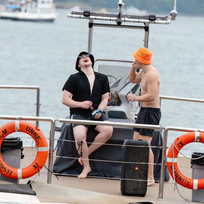 Two men are on a boating trip. One man, standing, wears an orange bucket hat and black swim shorts, holding a drink. The other man, sitting, sports a black hat and shirt while laughing exuberantly. The picturesque sea and other boats are visible in the background.