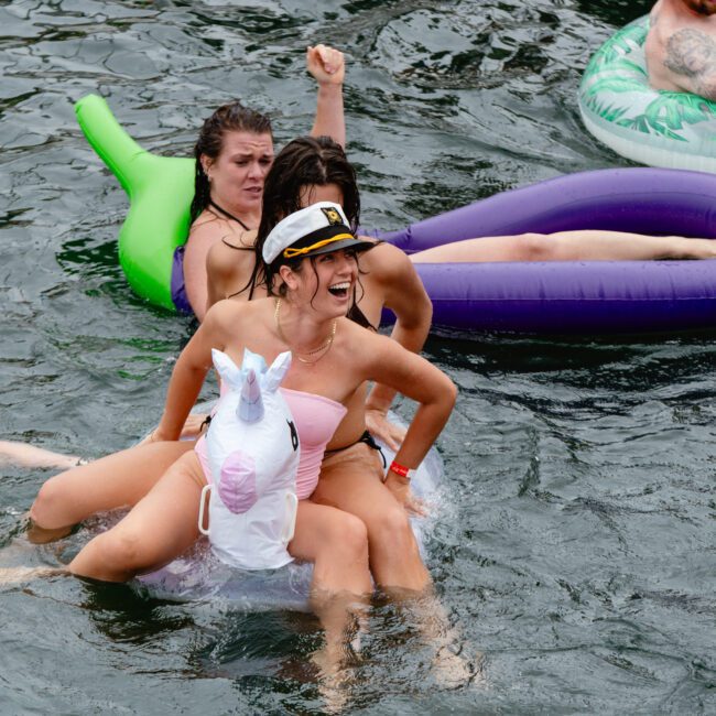 A woman in a captain's hat and a white one-piece swimsuit sits on an inflatable unicorn in a body of water, laughing with friends around her. Other people on inflatables and enjoying the water can be seen in the background.