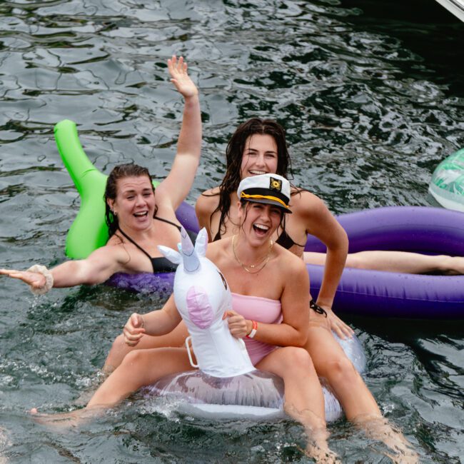 A group of people enjoy a fun time in the water. Four women, some on inflatable toys including a colorful unicorn, wave and smile at the camera. A man floats on a lounge float nearby. They all look relaxed and cheerful in the sunny weather, embracing the joyful moments together.