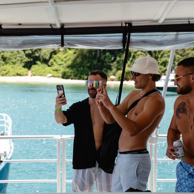 A group of three men stands on a boat taking a selfie. They are dressed in casual summer attire, including shorts and sunglasses, and they seem to be enjoying a sunny day near the water. Trees and another boat can be seen in the scenic background.
