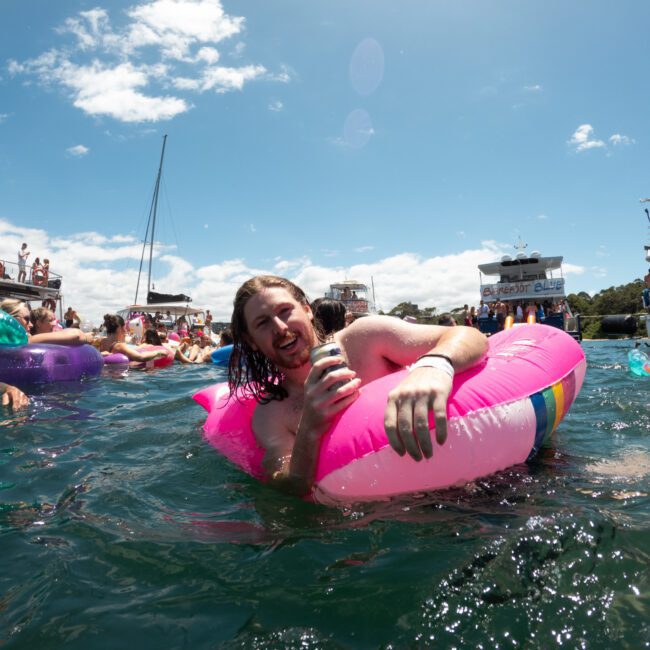 A person with long dark hair is enjoying a milkshake while relaxing in a pink inflatable on the water. Other people on inflatable floats and boats are in the background. The scene is bright and sunny, suggesting a fun, lively atmosphere on the water.