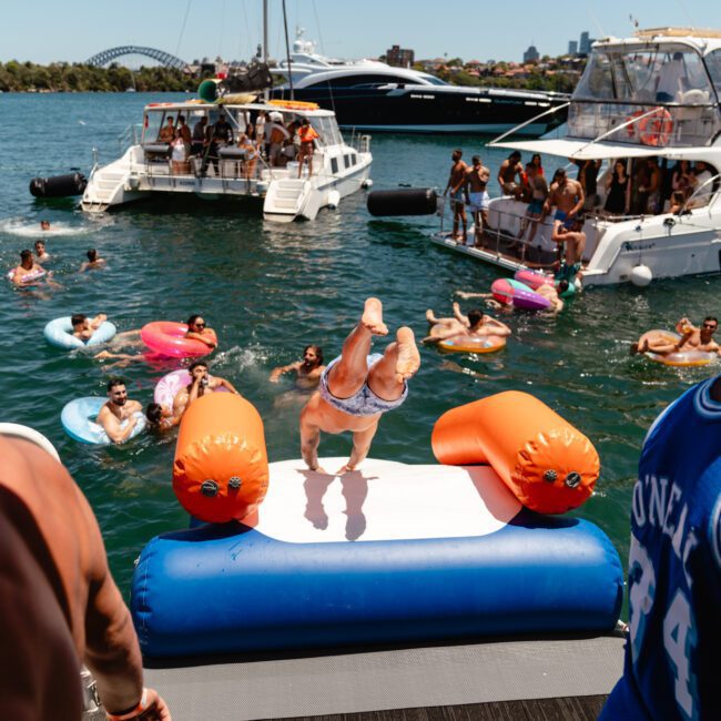 A lively scene on the water with people celebrating on multiple boats. In the center, a person is jumping into the refreshing water from an inflatable platform while others relax on colorful pool floats, creating a festive atmosphere. The background showcases a bridge and city skyline.