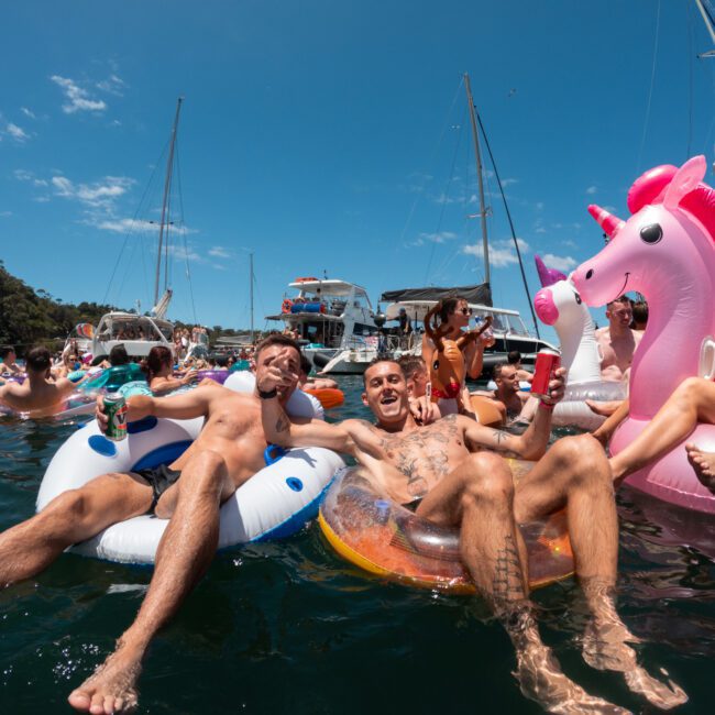 A lively scene of people enjoying a sunny day on the water. Two men are floating on inflatable rings, one on a donut and the other on a blue ring, drinking from cans. A child is playing nearby with an adorable pink unicorn float, surrounded by others having fun on various inflatables. Boats are in the background.