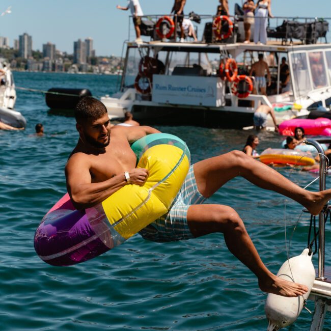 A man in swim shorts is slipping off a boat's ladder and falling into the water while holding a colorful inflatable ring. Several people are enjoying a sunny day on the boat, with city buildings visible in the distant background.