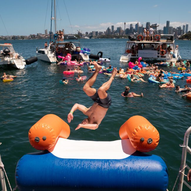 A person is mid-flip off an inflatable slide on a boat, surrounded by people on colorful floats in the water. Boats and a city skyline are visible in the background, creating an exhilarating scene perfect for a Sydney boat party hire.