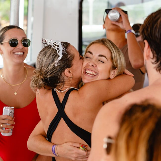 A group of people on a boat, smiling and enjoying a party. Two women are hugging in the center; one wears a tiara, and the other has long blonde hair. People around them are wearing swimsuits and sunglasses, holding drinks against the backdrop of sparkling water. The atmosphere is joyful and festive.