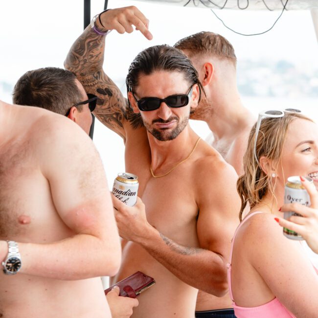 A group of people socializing outdoors, some without shirts. A man in sunglasses and a tattoo on his left arm is holding a can and pointing upwards. A woman in a pink bikini with a drink in hand stands nearby, while others mingle in the background at the lively gathering.