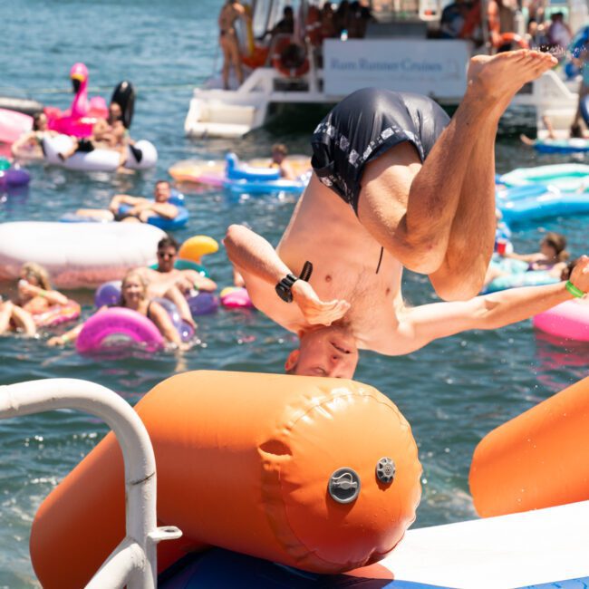 A man is mid-air in a headstand dive into a lake filled with people on inflatables during a sunny day. A luxury yacht hire Sydney is visible in the background.