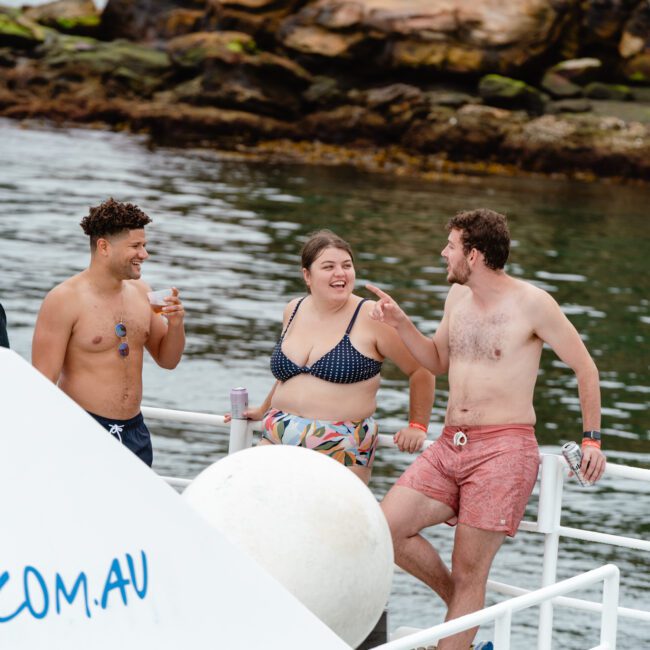 A group of people in swimwear stand and lean against a boat railing, chatting and laughing. They are on a boat near rocky shoreline waters. The warm, sunny weather enhances the cheerful ambiance. One person is wearing a hat, while another holds a refreshing drink.