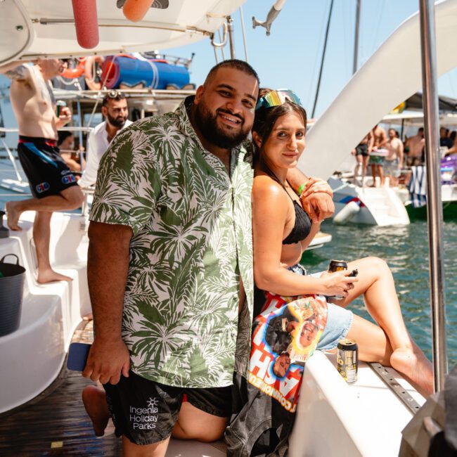 A man and a woman pose for a photo on the edge of a boat deck during a bright sunny day. The man wears a green tropical shirt and black shorts, while the woman in brown bikini bottoms and a yellow top holds a drink, with other boats and people creating an energetic ambiance in the background.