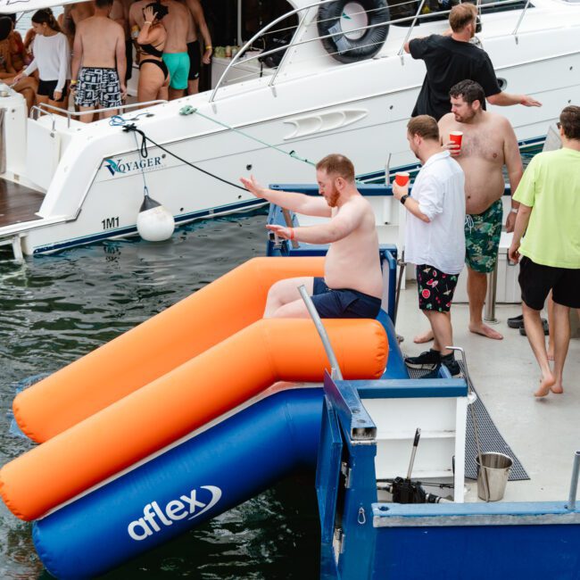 A group of people are gathered on a boat, some standing on the deck, others in swimwear near the water. One man is sitting on an inflatable water slide attached to the boat, preparing to slide into the lake. Several boats and people are visible in the background enjoying a sunny day.