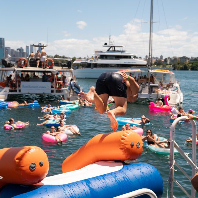 Person jumping off a boat's inflatable slide into the water, surrounded by people on colorful floaties, with other boats in the background on a sunny day during a Catamaran party Sydney.