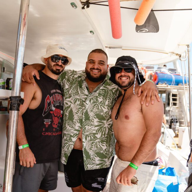 Three men are posing together aboard a boat, smiling towards the camera. The man in the middle is wearing a green tropical shirt and has his arms around the other two men, who are dressed in casual summer attire. A picturesque view of the water and distant shoreline is visible in the background.