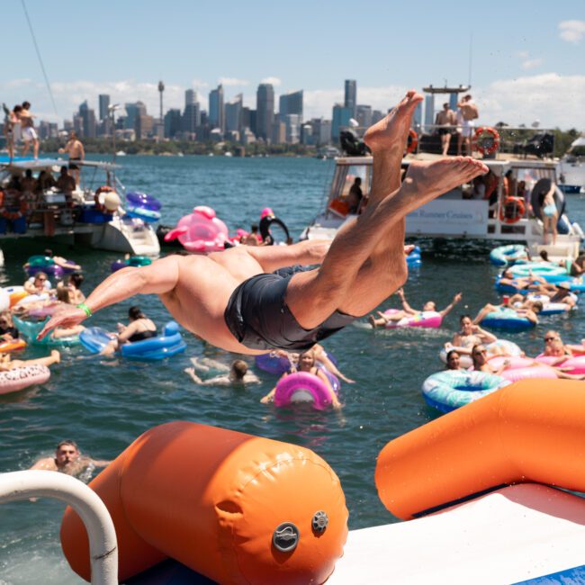 A man is jumping from an inflatable structure into the water, surrounded by people on floaties, with a city skyline in the background. It’s reminiscent of a fun-filled Sydney boat party hire, perfect for those looking to enjoy corporate boat events in style.