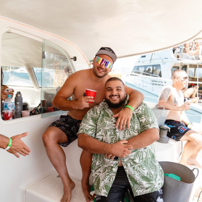 Two men are smiling on a boat. One is sitting while the other stands close behind, resting an arm around his shoulder. The seated man is holding a red cup. Both are wearing casual clothes, with others visible in the background. The "Sail Gustavo" logo can be seen at the bottom left corner of the image.