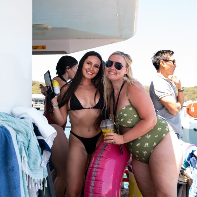 Two women in swimsuits and sunglasses pose together on a luxury yacht hire Sydney, one holding a drink and the other holding a pink inflatable. Other people can be seen in the background. Towels and bags are visible on the side.