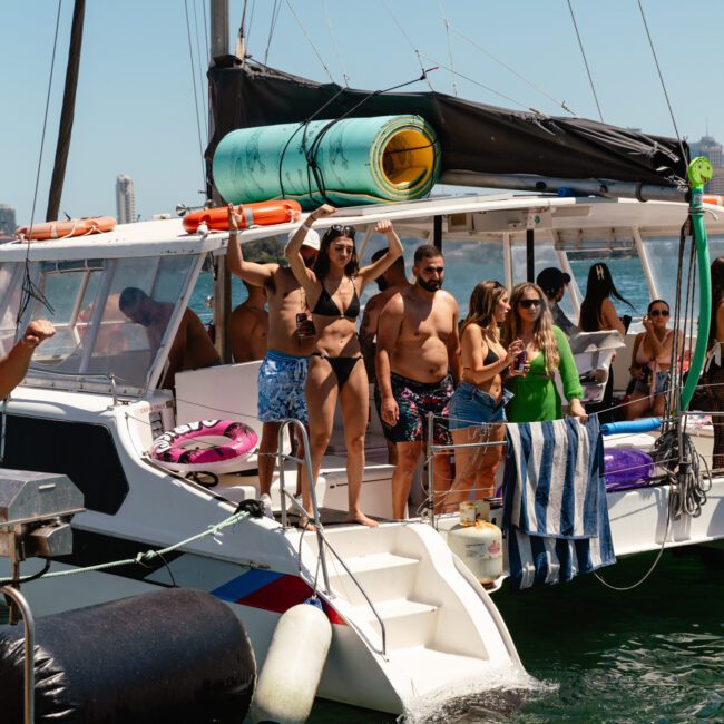 A group of people in swimwear stand and sit on a yacht with a white hull and black trim, moored in calm waters. The yacht has a stack of green and yellow floatation devices on top. Towels are draped over the railing, and the clear sky reveals some distant city buildings.