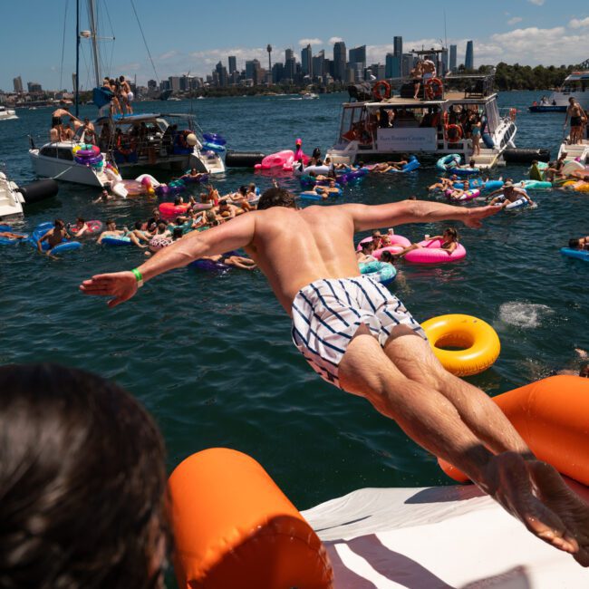 A man in striped swim trunks dives off an inflatable platform into the water, surrounded by boats and numerous people on colorful inflatables. A city skyline is visible in the background, hinting at a lively Catamaran party Sydney experience.
