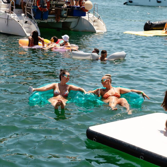 People enjoy a sunny day on the water near boats. Two individuals float on inflatable blue loungers, while others relax on inflatables and a floating platform. A woman on the right holds a phone, likely taking a photo. The festive and relaxed atmosphere is perfect for capturing memories.