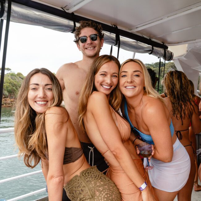 A group of friends enjoying a boat party. Three women in swimwear are smiling and posing for the camera, while a man in sunglasses stands behind them with his arm around one of the women. The picturesque background features sparkling water and a lush shoreline with trees.